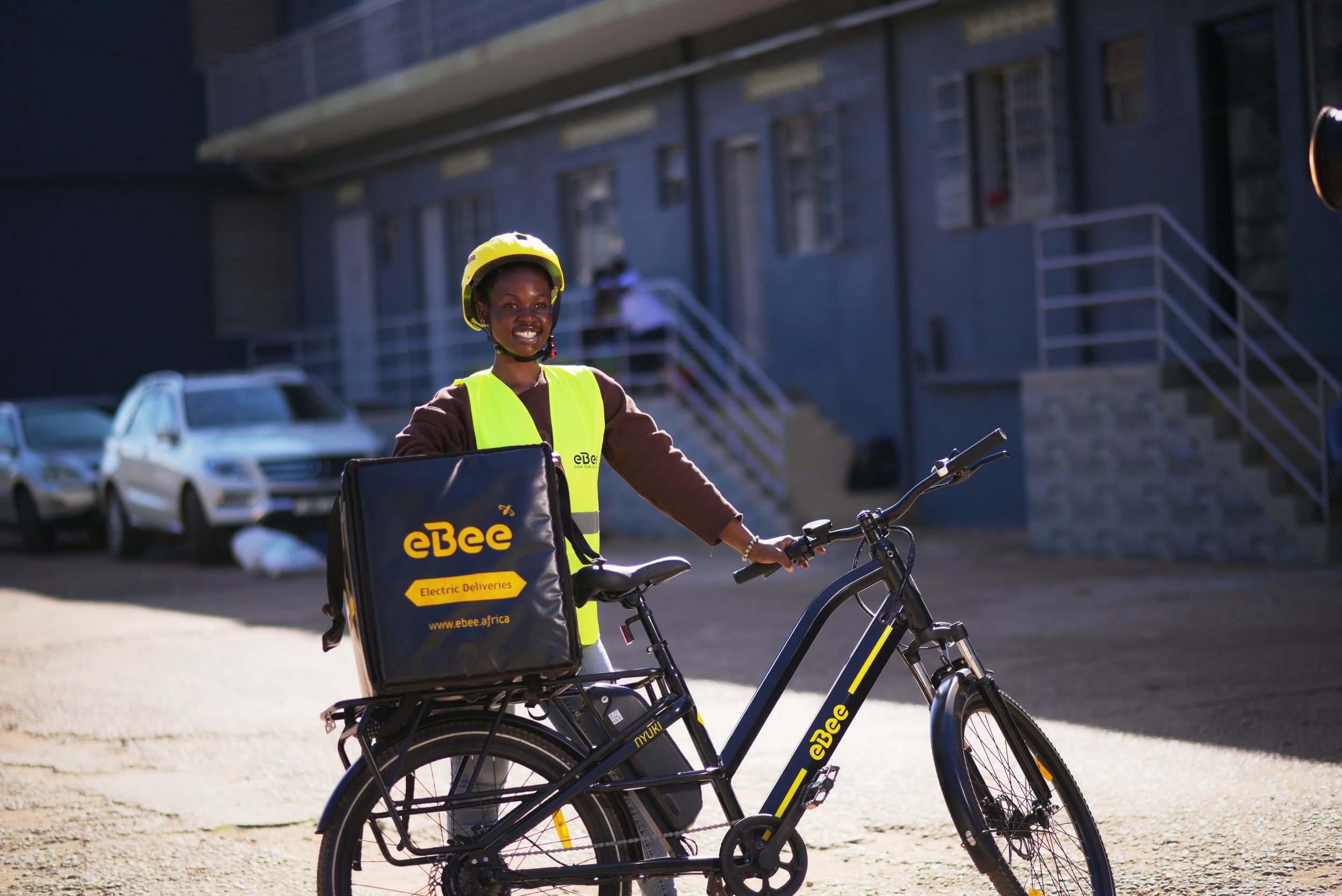 A smiling woman wearing a yellow helmet and reflective vest standing next to an e-bike with a delivery bag labeled 'eBee' in an urban setting